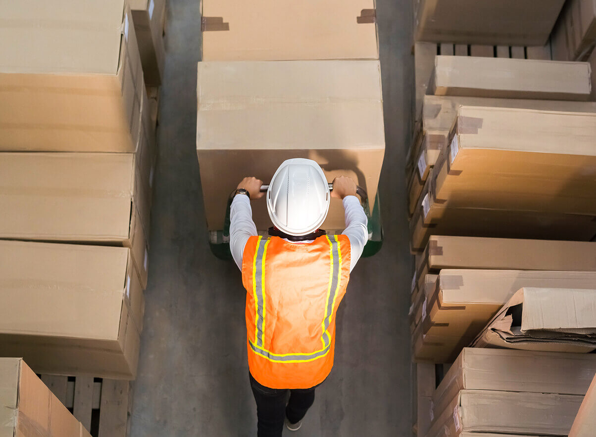 Bovenaanzicht van een medewerker met veiligheidshelm en reflecterend vest die een palletwagen met dozen verplaatst tijdens een magazijnontruiming.